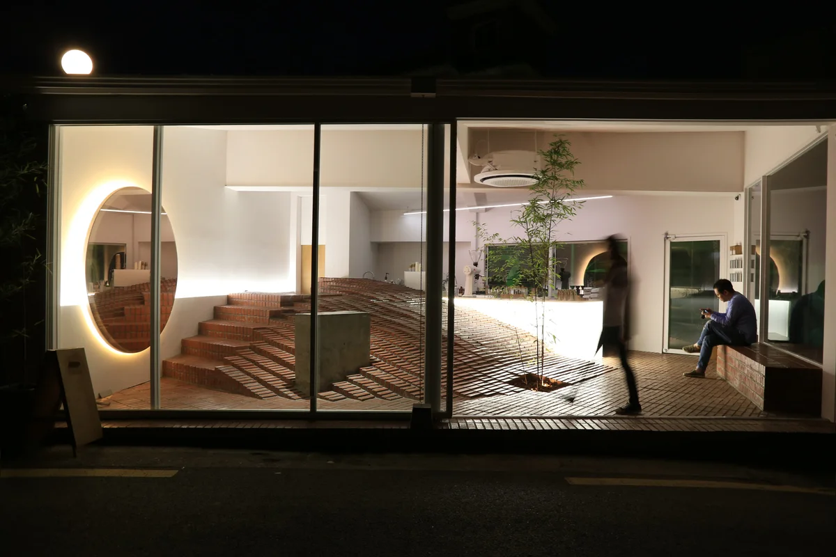 Coffee Nap’s Yeonnam glass frontage at night, with its terraced red-brick seating, circular mirror and bamboo tree visible inside.