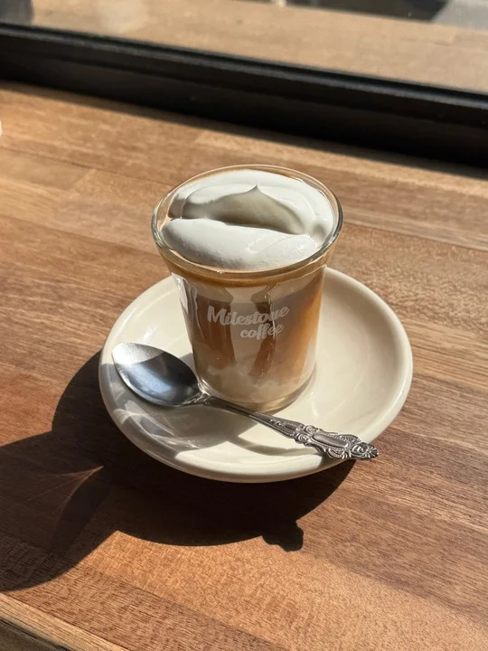 Cream-topped coffee in a Milestone-branded glass on a sunlit wooden window counter at the Hannam branch.